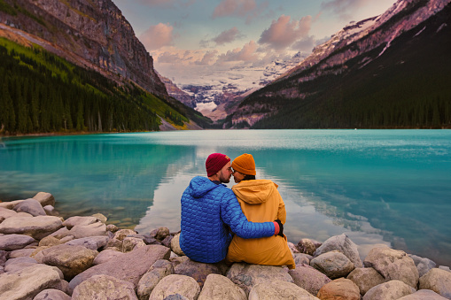 Lake Louise Banff National Park in the Canadian Rocky Mountains. A young couple of men and women sitting on a rock by the lake during a cold day in Autumn in Canada watching the sunset at the lake