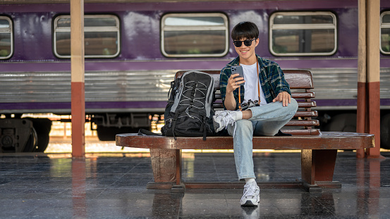 A young Asian male traveler is using his smartphone while sitting at a bench and waiting for his train at a railway station. solo traveler, backpacker, tourist