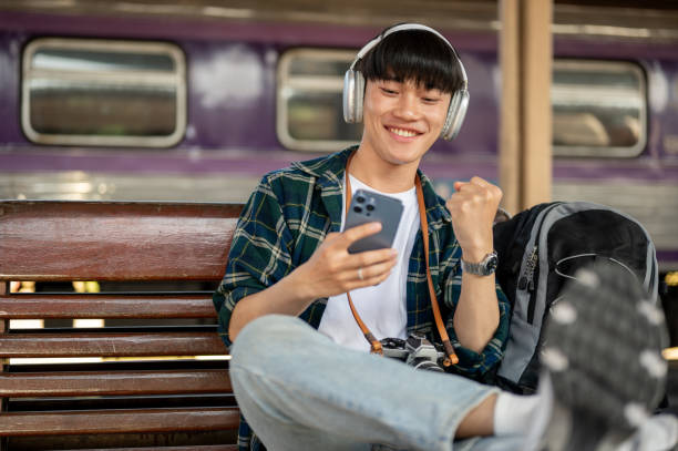 istock A cheerful Asian man backpacker watching something online on his phone while waiting for his train. 1933776824