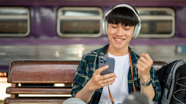 istock A cheerful Asian man backpacker watching something online on his phone while waiting for his train. 1933782121