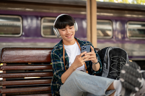 A happy and carefree Asian man backpacker is listening to music on his headphones and using his smartphone while sitting at a bench on a platform of a railway station, waiting for his train.