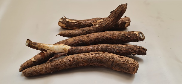 Five stalks of raw cassava on a beige background. The skin of the cassava is brown and still has soil attached to it. Before cooking, the singlong skin must be peeled first.