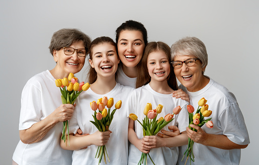 Happy women's day! Children daughters are congratulating mom and grandmothers giving them flowers tulips. Grannys, mom and girls smiling on light grey background. Family holiday and togetherness.