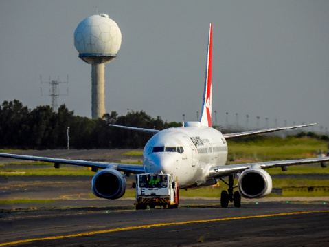 A Qantas Boeing B737-838 plane, registration VH-XZH, is being towed from the international terminal to the domestic terminal after arriving as flight QF122 from Queenstown.  She is scheduled to depart to Melbourne as flight QF487 in about two hours.  In the background is the airport's weather measuring structure, and navigational landing structures for the third runway. This image was taken on 13 January 2024 from Nigel Love Bridge, Mascot on a hot and sunny afternoon, with summer storm clouds starting to form.