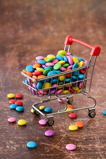 Stock photo showing close-up view of a heap of rainbow coloured smarties, candy coated chocolate drop sweet confectionary in a miniature, model shopping trolley. Unhealthy eating concept.