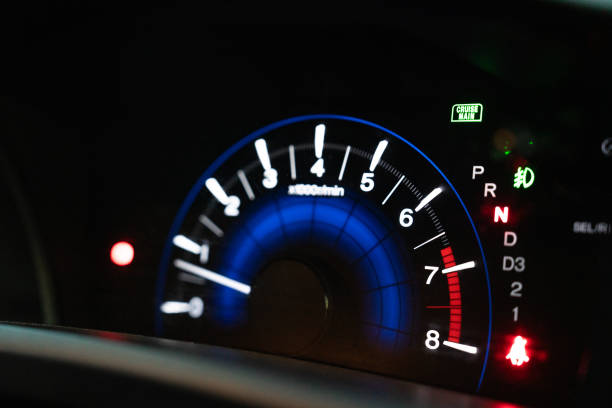 Angled close-up of a car speedometer glowing in red light, with the needle pointing near zero. The odometer and warning lights are partially visible, highlighting modern vehicle instrumentation.