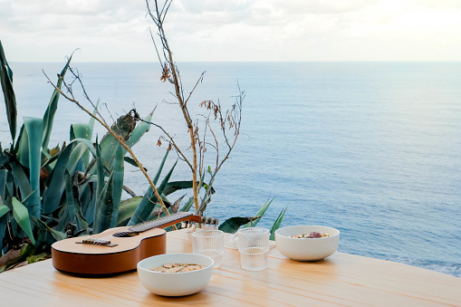 A peaceful setting featuring a guitar and refreshments on a wooden table, set against the backdrop of a calm ocean under soft daylight