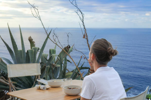 istock Unrecognizable young man Seated at a Wooden Table with Breakfast Set, Overlooking beautiful Ocean Horizon, Surrounded by Nature 1934123960