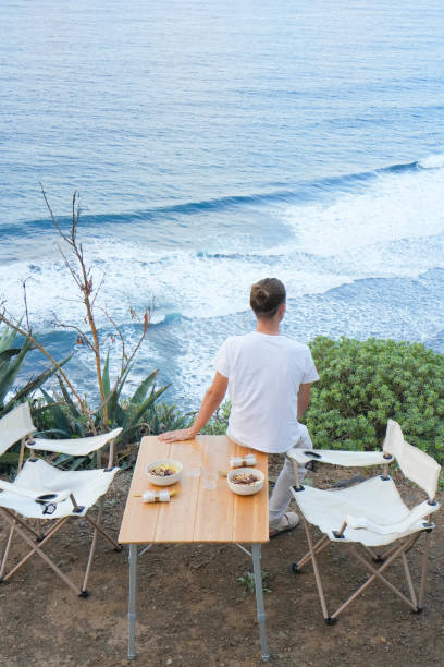 Dusk Serenity: Unrecognizable Solitary Man in White Shirt at Cliffside Dining Setup Overlooking the beautiful Ocean Waves