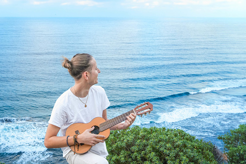 Side view of positive young musician in white casual clothes standing and playing acoustic guitar on shore of wavy blue sea