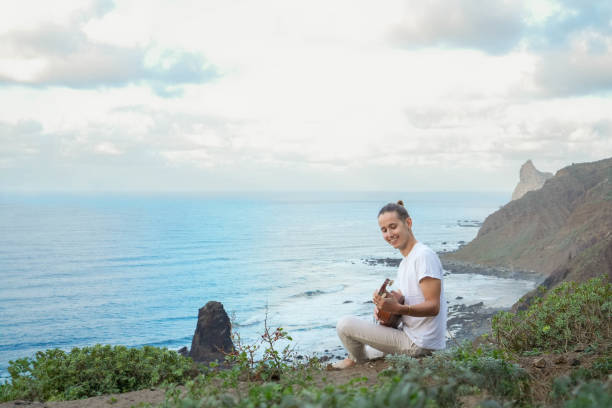 istock Young alternative man in White outfit on a Cliff, playing music in a ukelele while Overlooking the Vast Blue Sea Under a Clear Sky, Captured in a Vertical Frame 1934143483