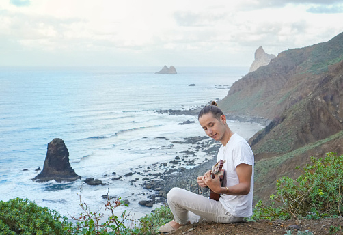 A tranquil moment of music amidst nature; young man serenades the rhythmic ocean waves with a ukulele
