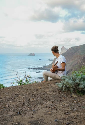 A tranquil scene of face less young man engrossed in playing the guitar, seated on a cliff with the vast ocean and rocky outcrops in the backdrop