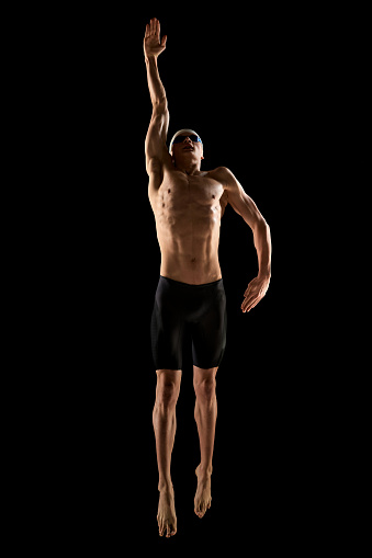 Male swimmer with highlighted muscle definition, poised in swimming stance. Athletic young man, swimmer showing swimming techniques over black background. Concept of aquatic sports, competition