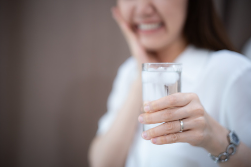 Focus on a cold glass of water with blur young Asian woman feel sensitive on her tooth face, Lady touch at her chin with sensitive teeth expression, with brown curtain background.