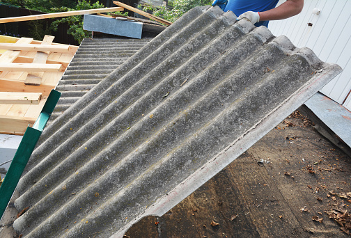 A building contractor is removing old harmful and dangerous for health corrugated asbestos roof tile. Repair house asbestos roof. Roofing construction.