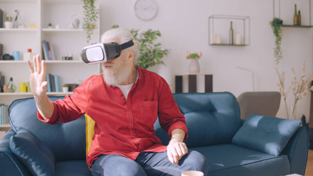 A mature man experiences virtual reality, seated on a sofa with a VR headset, as he reaches out into virtual space