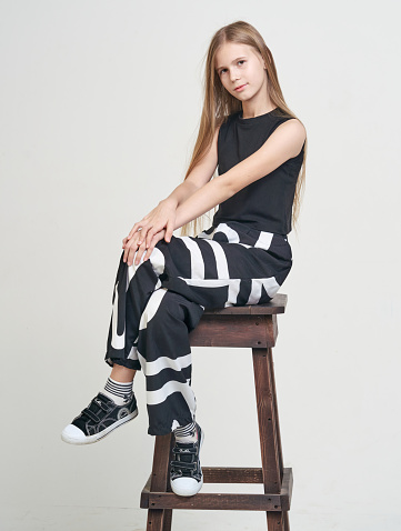 Beautiful blond-hair preteen girl dressed in a top and in black pant. She is sitting with cross legs on a bar stool, looking at the camera. Full length portrait. Studio shot, white background