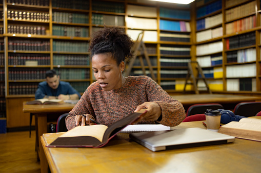 African American college student going through book while doing her research in a library.
