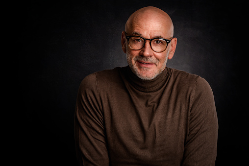 Portrait of mature man with glasses smiling to camera against isolated black background. Confident male wearing turtleneck sweater.