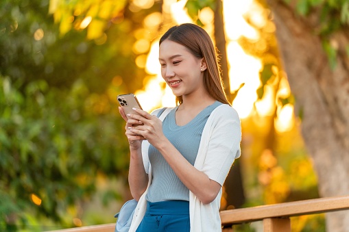 Gen Z Asian Woman Using Phone under a Tree Against Beautiful Sunset in a Public Park