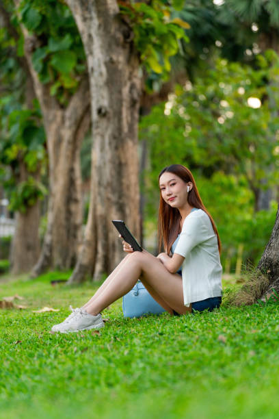 istock Gen Z Asian Woman Using Earbuds and Digital Tablet under a Tree in a Public Park 1934404720