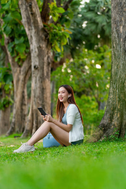 istock Gen Z Asian Woman Using Earbuds and Digital Tablet under a Tree in a Public Park 1934405257