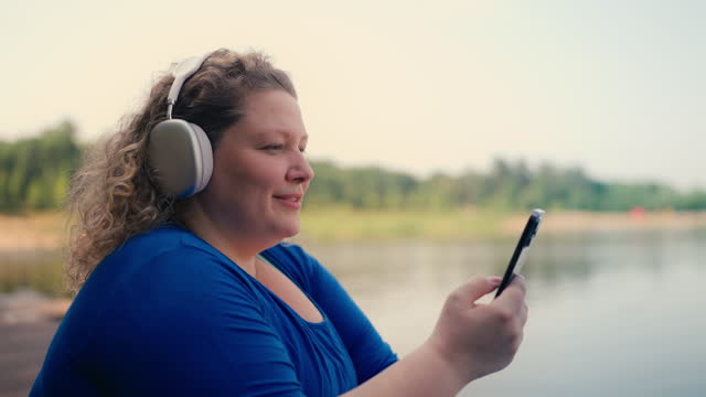 Plus size woman listening music on earphones, using mobile player app on gadget