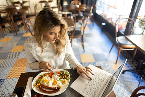 A beautiful mid adult blond woman, sitting in a cafe, she is eating eggs and sausages. There is a laptop in front of her, she is typing on it.