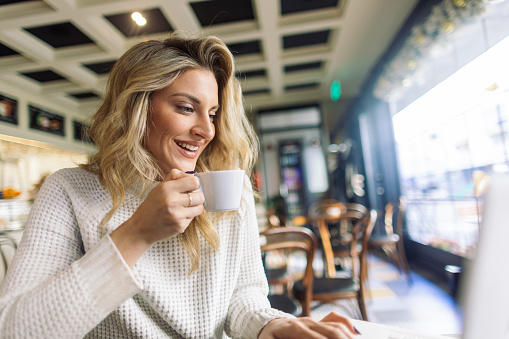 A beautiful mid adult blond woman, sitting in a cafe, typing on her laptop and drinking coffee. She is maybe having a video call, she is smiling, looking happy.