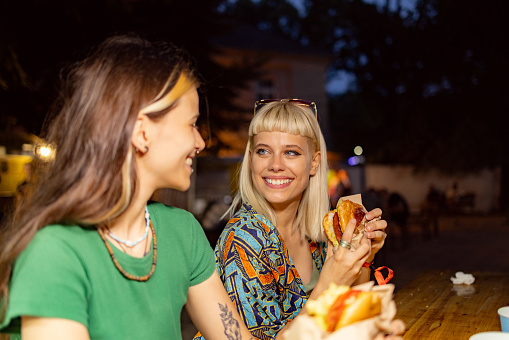 Young happy woman eating burger while communicating with her friend during a music festival by night.
