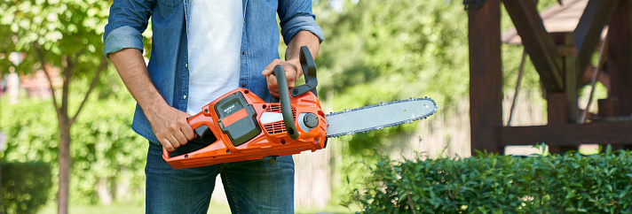 Unrecognizable craftsman standing with cordless chain saw, working in summer garden. Crop view of strong handyman in denim clothes holding modern electric saw. Concept of garden equipment.