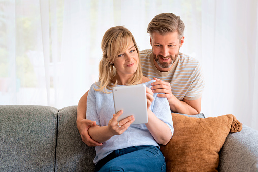 Happy couple relaxing on the sofa at home. Attractive mid aged woman and handsome man using digital tablet and browisng on the internet or having video call.