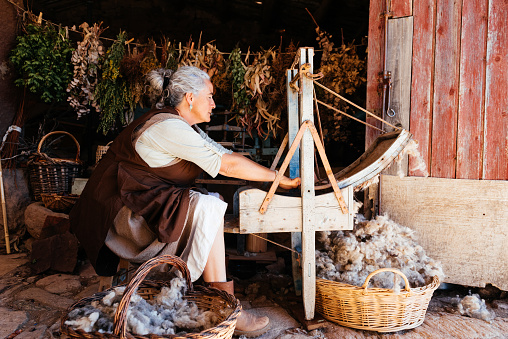 Side view of senior female in casual farm clothes sitting with baskets of fresh wool and while working and threshing wool using a carding machine in local cloth making shop