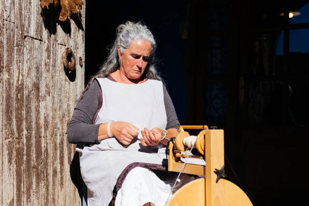 Focused senior female artisan in casual clothes using old thread maker spinning wooden machine for making thread while working at against grungy wall