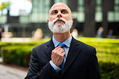 Mature bald stylish business man portrait with a white beard outdoor adjusting his necktie