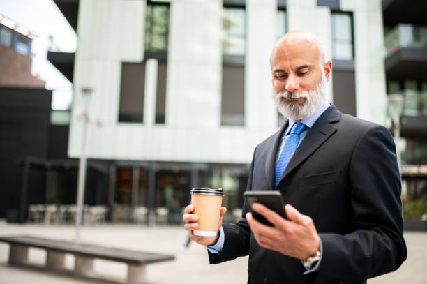 istock Stylish senior bald manager whit white beard using his smartphone outdoor while drinking a coffee 1934475643