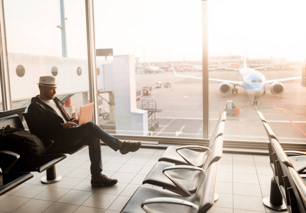 istock Airport Terminal: Businessman Uses Laptop Computer, Waiting for His Flight. Traveling Entrepreneur Remote Work Online Sitting in Boarding Lounge of Airline Hub with Airplanes Arriving, 1934492436