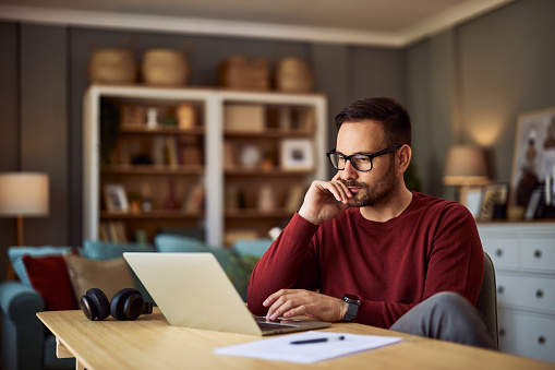 A busy, focused young adult male freelancer sitting at a desk and resting his head on his hand while working on a project on his laptop.