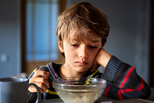 Upset European male child having breakfast while sitting in front of  transparent bowl of porridge in dimly lit kitchen with gray walls.