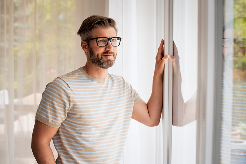 Mid aged man standing at the window and looking out. Handsome male having grey hair and wearing glasses.