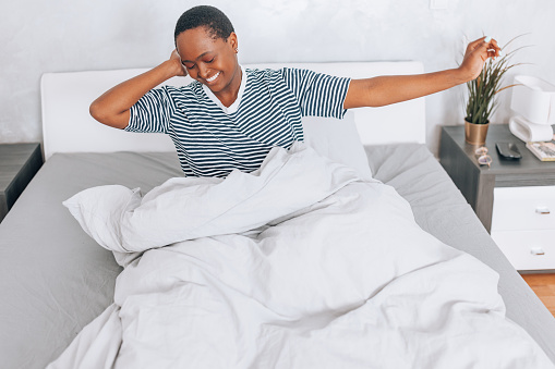 This image shows a young woman, stretching while sitting in bed, her gaze looking away in thought, encapsulating the calmness and relaxation of waking up at home