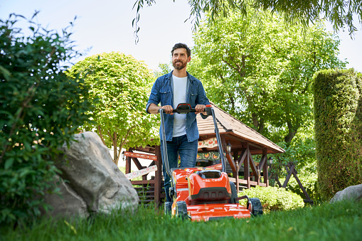 Smiling guy with beard trimming overgrown green lawn with electric mower in garden. Low angle view of happy male gardener in shirt using lawn trimmer, while landscaping. Concept of seasonal work.