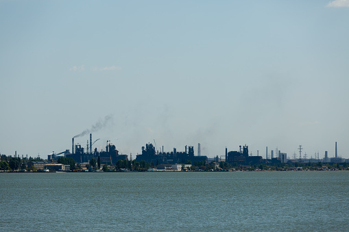 Smoke rises against blue sky, signaling pollution. Industrial skyline across water with emission from stacks at metallurgical plant. Manufacturing complex emits byproduct, symbolizing eco issues.