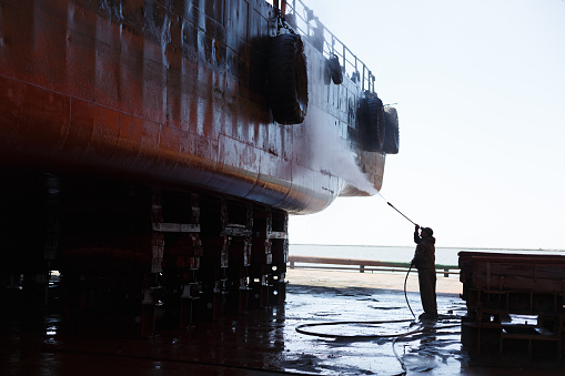 Ship maintenance, maritime industry. Worker cleans vessel hull in dry dock with high-pressure washer. Man in protective gear, silhouetted against vessel. Industrial cleaning, shipping repairs.