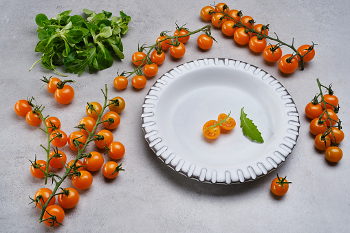 Sliced yellow cherry tomato and leaf arugula in a white porcelain bowl on the gray granite table. Sprig of cherry tomatoes with green salad on a gray concrete background. Selective focus. Flat lay