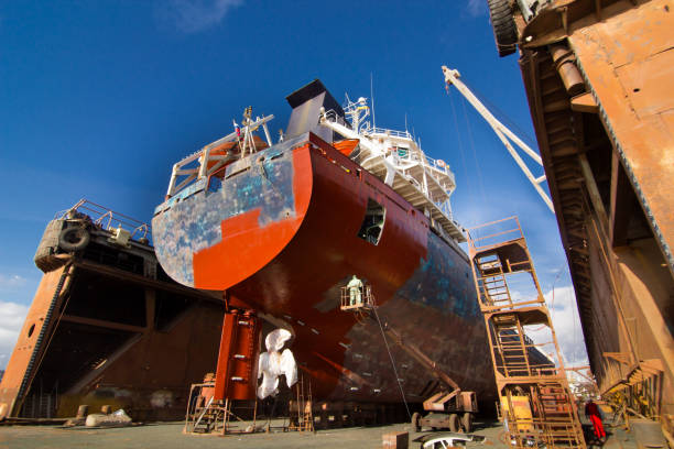 istock Commercial vessel undergoes maintenance in floating dock. Workers paint, repair hull, under clear skies at shipyard. Maritime industry, vessel refurbishment, drydock operation captured in daylight. 1934560882