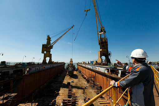 Maritime worker points, directs crane at shipyard under sunny sky. Engineer oversees vessel maintenance in dry dock. Industrial worker inspects, guides ship overhaul.
