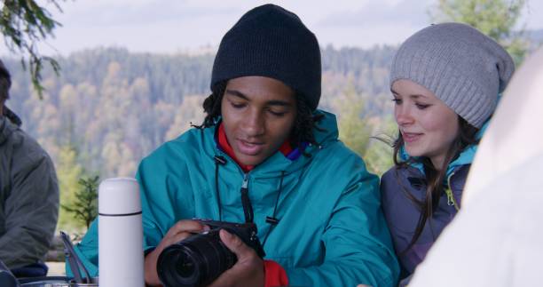 Diverse hiking buddies rest in wooden gazebo after long trek in forest. African American teenager smiles, talks and shows photos from camera to other tourists. Nature discovery and tourism concept.