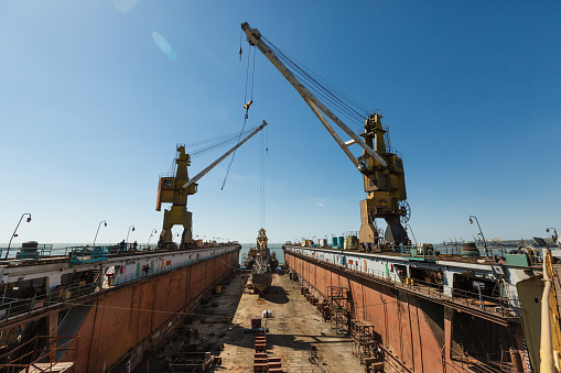 Maritime vessel reconstruction, heavy machinery operational. Ship repair dock with towering cranes, industrial setting under clear sky. Infrastructure for seafaring maintenance, nautical hub.